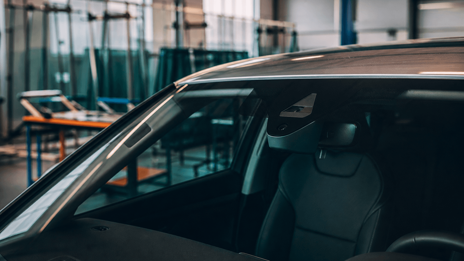 Windshield with a forward camera housing on a modern car in a glass workshop bay, soft daylight, no readable logos or plates.